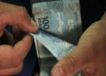 Mãos seguram e contam notas de 100 reais brasileiros em fundo escuro, representando os pagamentos do PIS/Pasep e do BPC de maio