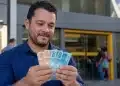 Homem segurando cédulas de real em frente a uma agência bancária, representando o saque do PIS/Pasep de junho.