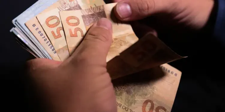 Mãos contam várias cédulas de cinquenta reais sobre fundo escuro.