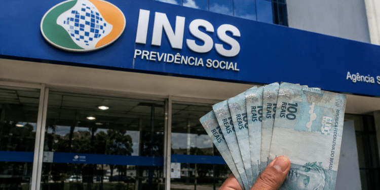 Mão segurando cédulas de cem reais em frente a uma agência do INSS, em referência ao pagamento de R$ 1.621 ou R$ 2.431,50 da segunda parcela do 13º salário em 2026.