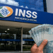 Mão segurando cédulas de cem reais em frente a uma agência do INSS, em referência ao pagamento de R$ 1.621 ou R$ 2.431,50 da segunda parcela do 13º salário em 2026.