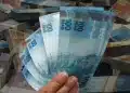 Mão segurando notas de R$ 100, com maços de dinheiro ao fundo, ilustrando os pagamentos de R$ 152,99, R$ 178,48 e R$ 229,48 liberados para CPFs com finais de 0 a 9.