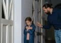 Homem adulto apontando dedo para menina ruiva de braços cruzados em postura defensiva, em corredor residencial, simbolizando o narcisismo parental.
