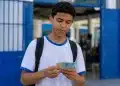 Estudante segurando notas de real na entrada de escola pública, representando os últimos a receber os pagamentos do Pé-de-Meia de maio.