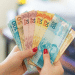 Mãos com unhas vermelhas exibem notas de 50, 10, 20 e 100 reais em formato de leque, mostrando a segunda família do real, simbolizando o pagamento de R$ 4.116.