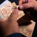 Mãos masculinas contando várias cédulas de R$ 50 do Banco Central do Brasil contra fundo escuro, simbolizando o pagamento de R$ 1.486 do PIS/Pasep 2026.