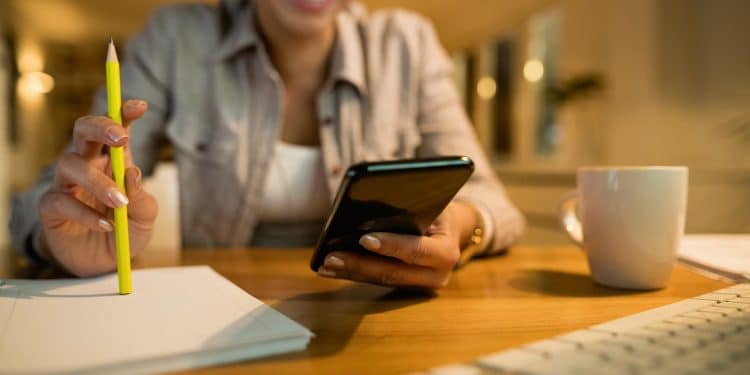 Mulher sorridente segura o celular em uma das mãos e um lápis na outra diante de caderno aberto e teclado sobre mesa de madeira