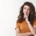 Jovem mulher de cabelo cacheado e camiseta laranja com a mão no rosto em expressão pensativa sobre fundo branco