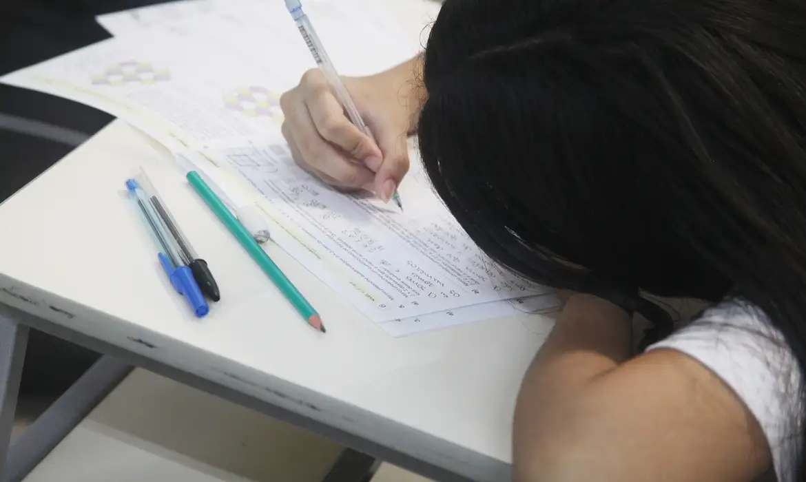 Jovem estudante escreve em folha de prova sobre carteira escolar enquanto se prepara para a redação do Enem 2026
