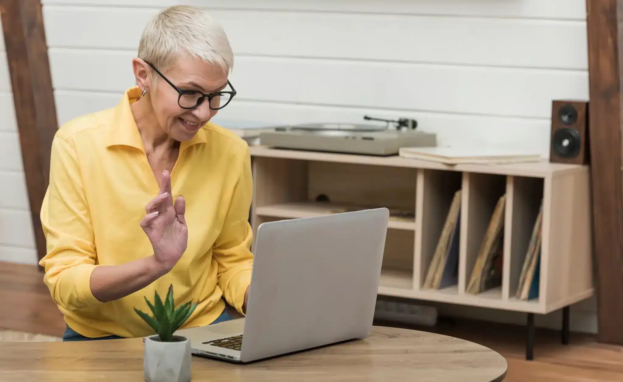 Mulher de meia-idade com camisa amarela usa notebook em videochamada em ambiente doméstico, exemplo de profissional que se prepara continuamente para as mudanças no mercado de trabalho.