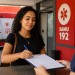 Mulher de cabelos cacheados preenche ficha com caneta azul enquanto atendente de uniforme azul segura prancheta em unidade do SAMU 192.