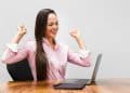 Mulher de camisa rosa comemora em frente ao notebook, simbolizando as profissões com salários de R$ 7 mil.