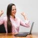 Mulher de camisa rosa comemora em frente ao notebook, simbolizando as profissões com salários de R$ 7 mil.