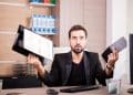 Homem de barba com blazer preto segura prancheta com gráficos na mão direita e tablet na esquerda, com expressão de surpresa, simbolizando as profissões em que faltam profissionais.
