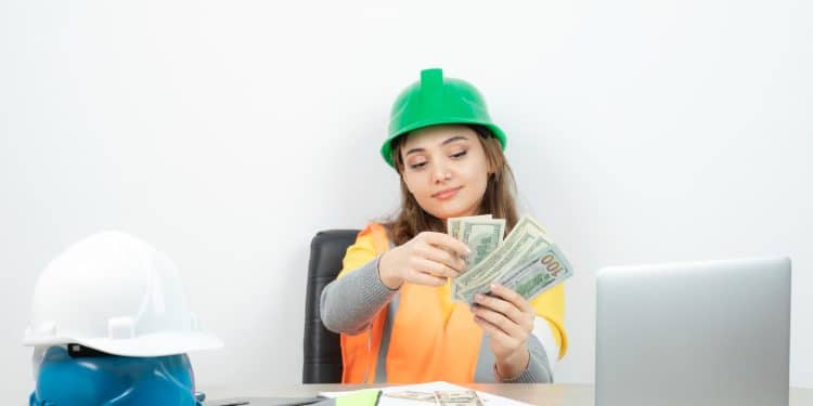Mulher com capacete verde e colete laranja conta cédulas de dinheiro sentada a uma mesa com notebook e capacetes de obra