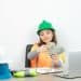 Mulher com capacete verde e colete laranja conta cédulas de dinheiro sentada a uma mesa com notebook e capacetes de obra