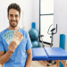 Profissional de saúde em scrub azul segura notas de 100 e 50 reais em clínica com maca e bicicleta ergométrica, simbolizando as opções renda extra para fisioterapeutas.