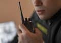 Vigilante de uniforme preto com inscrição amarela segura rádio comunicador próximo à boca durante ronda, simbolizando as opções de renda extra para vigilantes.