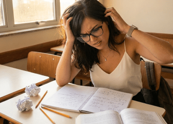 Estudante de óculos leva as mãos à cabeça diante de caderno com cálculos e papéis amassados sobre a carteira.