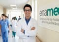 Médico de jaleco branco e óculos, de braços cruzados, sorri em corredor de hospital ao lado de banner do Enamed, o Exame Nacional de Avaliação da Formação Médica