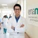 Médico de jaleco branco e óculos, de braços cruzados, sorri em corredor de hospital ao lado de banner do Enamed, o Exame Nacional de Avaliação da Formação Médica