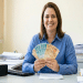 Mulher de camisa azul segura notas de R$100, R$50 e R$20 em leque, sorrindo em mesa de escritório com computador e documentos, simbolizando o salário de um auxiliar administrativo.