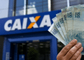 Mão segurando cédulas de cem reais em frente a uma agência da Caixa, em referência ao Saque Calamidade do FGTS de até R$ 6.220 liberado para novos municípios em 2026.