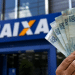 Mão segurando cédulas de cem reais em frente a uma agência da Caixa, em referência ao Saque Calamidade do FGTS de até R$ 6.220 liberado para novos municípios em 2026.