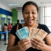 Mulher sorrindo segura cédulas de real dentro de agência bancária após sacar pagamento de até R$ 6.220 da Caixa