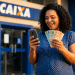 Mulher sorri em frente a uma agência da Caixa ao solicitar pelo celular o Saque Calamidade do FGTS e segura notas de R$ 100 e R$ 50