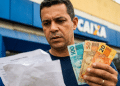 Homem confere papel em frente a uma agência da Caixa segurando notas de R$ 100, R$ 50 e R$ 20, em referência ao Saque Calamidade do FGTS.
