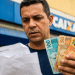 Homem confere papel em frente a uma agência da Caixa segurando notas de R$ 100, R$ 50 e R$ 20, em referência ao Saque Calamidade do FGTS.