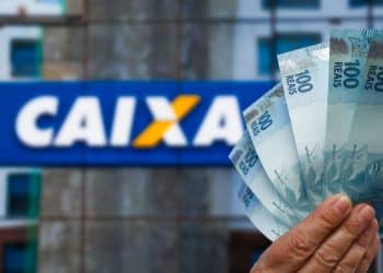 Mão segura cédulas de cem reais diante de fachada da Caixa Econômica Federal, em referência ao saque de até R$ 6.220