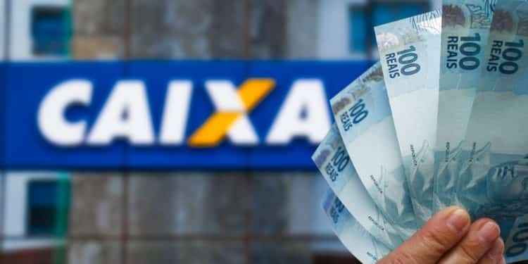 Mão segura cédulas de cem reais diante de fachada da Caixa Econômica Federal, em referência ao saque de até R$ 6.220
