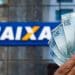 Mão segura cédulas de cem reais diante de fachada da Caixa Econômica Federal, em referência ao saque de até R$ 6.220