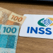 Cartão branco com logo INSS e bandeira do Brasil ao lado de notas de 100 reais e moedas sobre superfície de madeira, simbolizando a segunda parcela do 13º salário para quem recebe até R$ 1.621, atual salário mínimo.