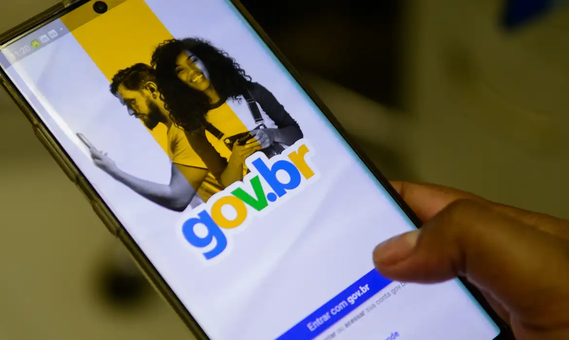 Mão segura smartphone com a tela de entrada do gov.br aberta exibindo imagem de dois usuários e a opção de acessar a conta