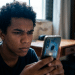 Jovem usa o celular com expressão concentrada e, ao redor, a escrivaninha e a estante de um quarto