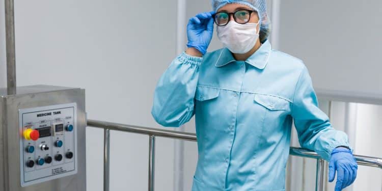 Profissional com jaleco azul, touca, máscara e luvas ajusta óculos próximo a painel de controle em sala limpa industrial, simbolizando a profissão de