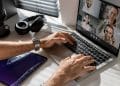 Mãos masculinas digitando em laptop prateado durante videoconferência com 4 colegas, headphone preto e tablet ao lado, simbolizando os trabalhos remotos de 2026 que pagam mais de R$ 8 mil.