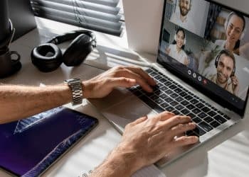 Mãos masculinas digitando em laptop prateado durante videoconferência com 4 colegas, headphone preto e tablet ao lado, simbolizando os trabalhos remotos de 2026 que pagam mais de R$ 8 mil.
