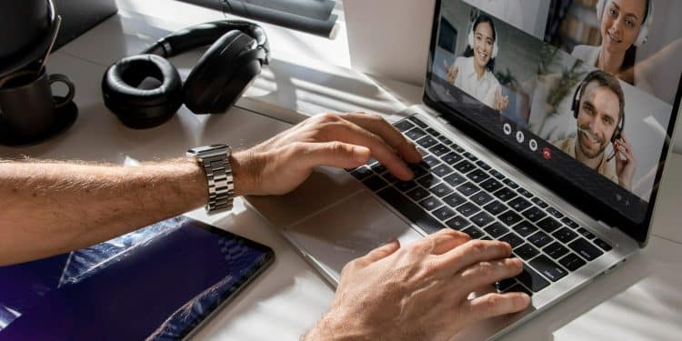 Mãos masculinas digitando em laptop prateado durante videoconferência com 4 colegas, headphone preto e tablet ao lado, simbolizando os trabalhos remotos de 2026 que pagam mais de R$ 8 mil.
