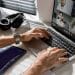 Mãos masculinas digitando em laptop prateado durante videoconferência com 4 colegas, headphone preto e tablet ao lado, simbolizando os trabalhos remotos de 2026 que pagam mais de R$ 8 mil.