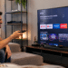 Homem de camiseta azul sentado no sofá aponta controle remoto para Smart TV com apps de streaming