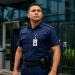 Vigilante com uniforme azul-marinho e crachá de identificação em frente a prédio comercial com fachada de vidro.