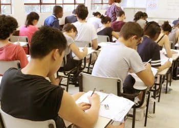 Sala de aula com diversos estudantes sentados em carteiras realizando uma prova com folhas e canetas