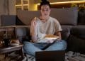 Mulher come batata frita enquanto assiste notebook em casa.