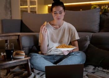 Mulher come batata frita enquanto assiste notebook em casa.