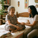 Idosa e mulher sorrindo juntas em quarto decorado com plantas e artesanato.