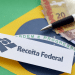 Papel com logo e nome Receita Federal sobre bandeira do Brasil, notas de real e caneta.